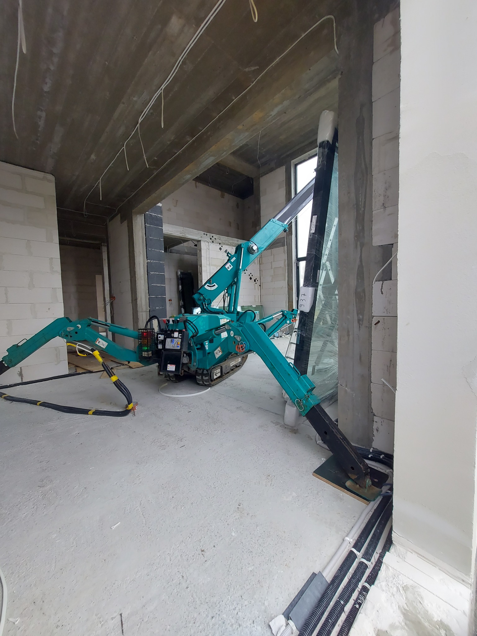 Mały, gąsienicowy dźwig w kolorze turkusowym ustawiony w surowym wnętrzu budynku w trakcie montażu okna. Widoczne przewody elektryczne i niedokończone ściany.