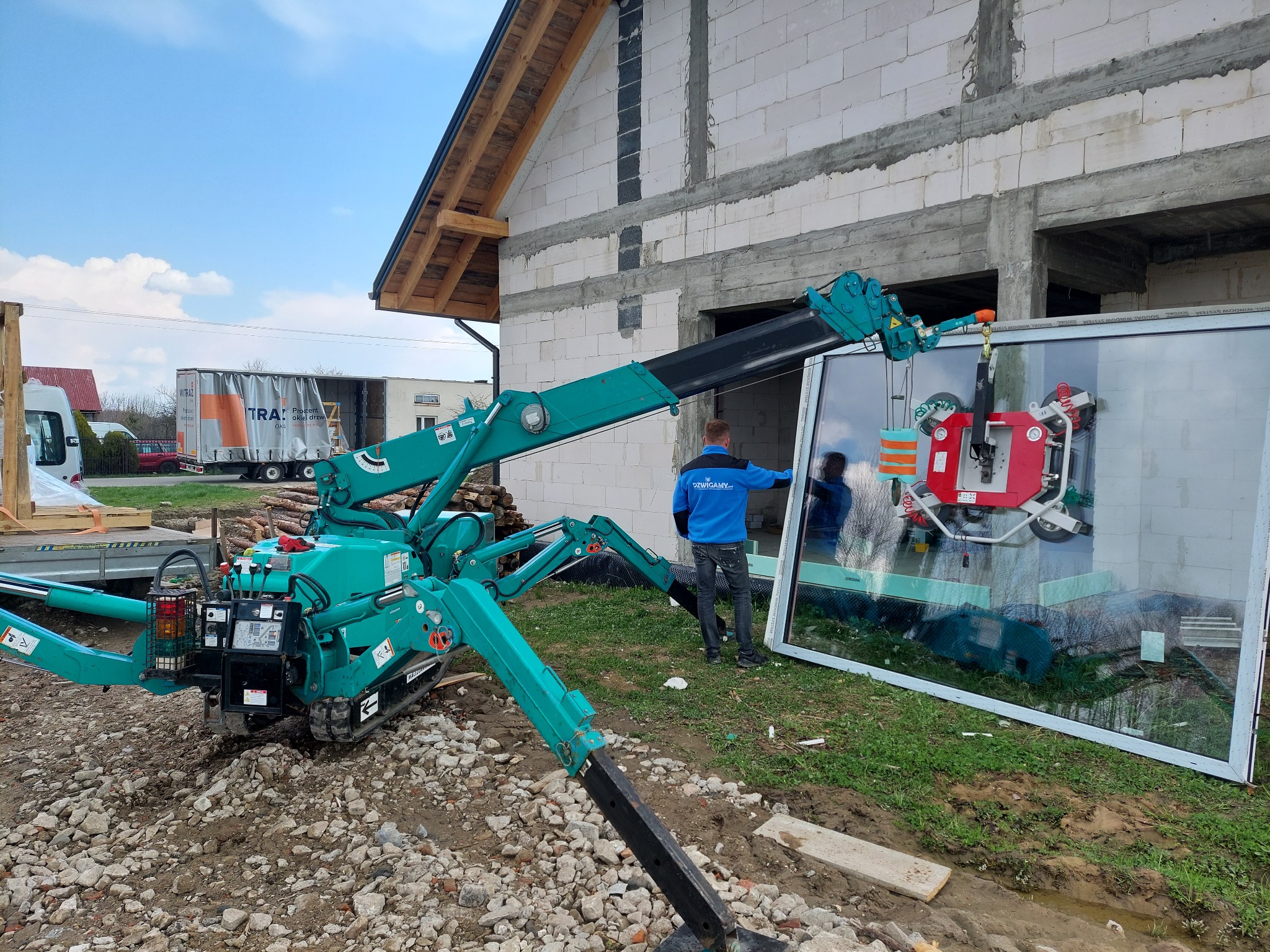 Kompaktowy dźwig pająk w kolorze turkusowym podnosi duże okno z ramą, widoczny pracownik w niebieskiej bluzie i budynek w trakcie budowy z białych pustaków.