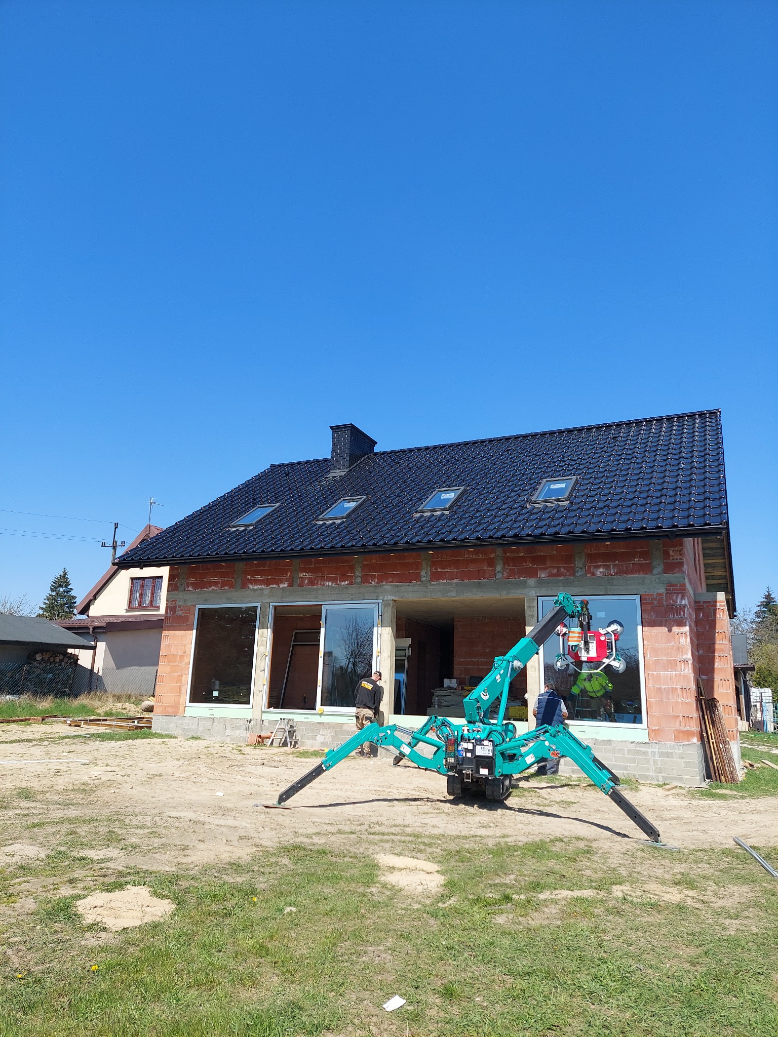 Turkusowy mini-dźwig pająk z przyssawkami podnosi dużą szybę okienną na placu budowy domu z czerwoną cegłą i czarnym dachem w słoneczny dzień.