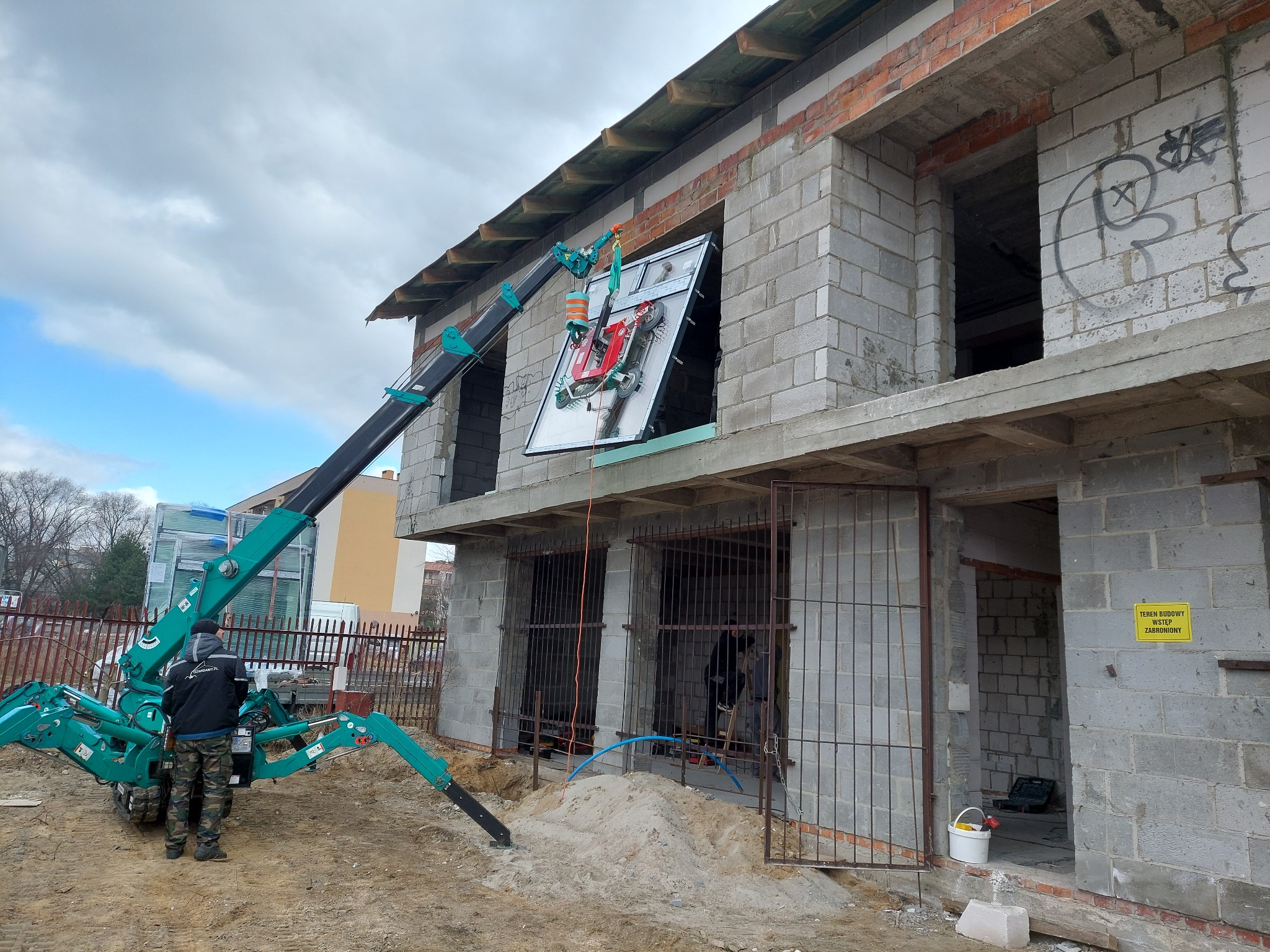 Kompaktowy dźwig z przyssawkami montuje duże okno w surowym stanie budynku z szarego betonu komórkowego.