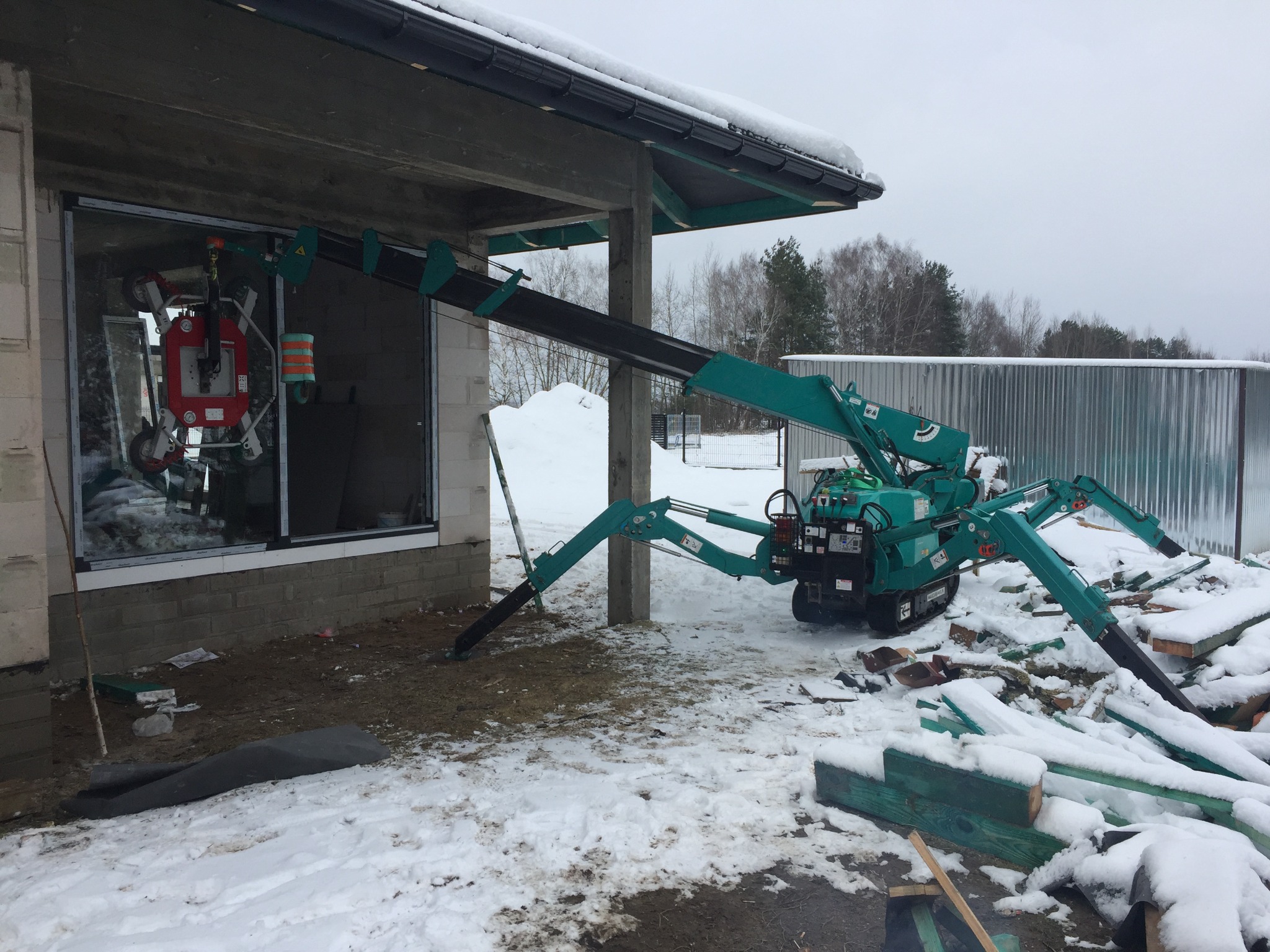 Mały, zielony dźwig pająk z przyssawkami montuje okno w surowym stanie budynku, na zewnątrz leży śnieg.