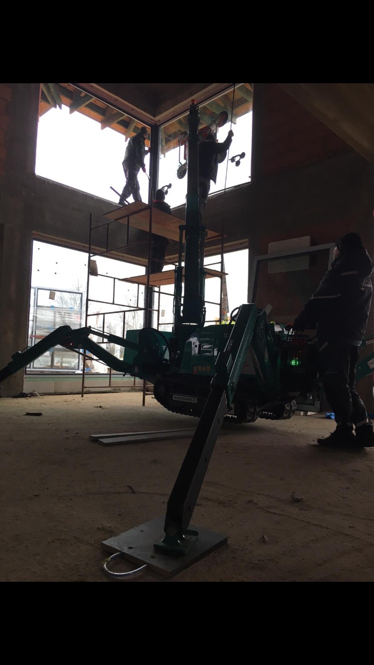 Montaż okien za pomocą małego dźwigu gąsienicowego w surowym stanie budynku, pracownicy na rusztowaniu w tle, widoczne przyssawki do szkła.