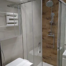 Nowoczesna kabina prysznicowa z przesuwnymi drzwiami, chromowana armatura, drewnopodobne płytki na ścianie, biały grzejnik drabinkowy i pralka w wykończonej łazience.