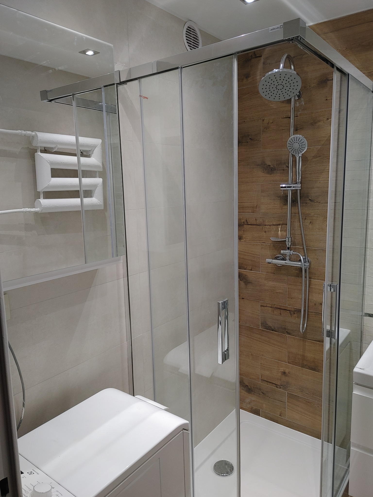 Nowoczesna kabina prysznicowa z przesuwnymi drzwiami, chromowana armatura, drewnopodobne płytki na ścianie, biały grzejnik drabinkowy i pralka w wykończonej łazience.