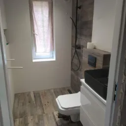 Nowoczesna łazienka z drewnopodobnymi płytkami na podłodze i ścianie, czarną armaturą i umywalką, widok zza drzwi.