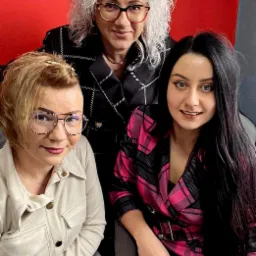 Portret trzech uśmiechniętych kobiet w różnym wieku i w stylizowanych marynarkach na czerwonym tle.