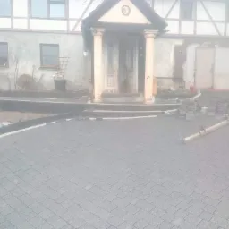 Ułożona szara kostka brukowa na podjeździe przed domem z kolumnami, widoczne niedokończone fragmenty i narzędzia.
