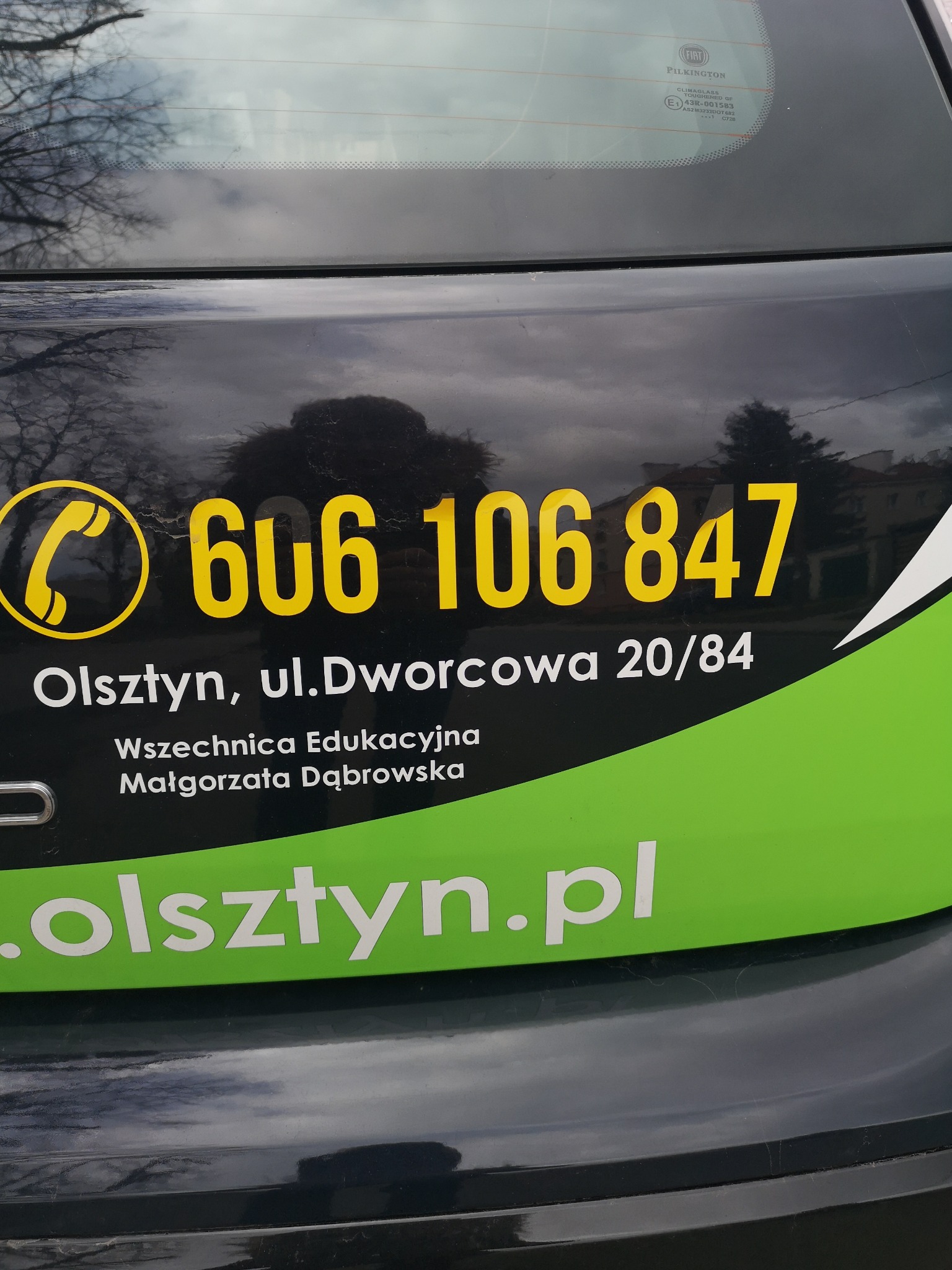 Reklama z numerem telefonu i adresem na tylnej klapie samochodu w Olsztynie, z nazwą Wszechnicy Edukacyjnej Małgorzaty Dąbrowskiej i adresem strony internetowej.