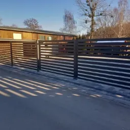 Nowoczesne, poziome, metalowe ogrodzenie posesji w kolorze grafitowym, z widocznymi cieniami rzucanymi przez konstrukcję na szarą kostkę brukową i asfalt.