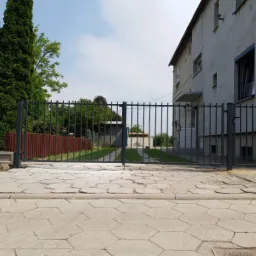 Nowoczesna, metalowa brama wjazdowa z pionowymi szczeblami, widok z poziomu chodnika, w tle dom z szarą elewacją i zielony ogród.