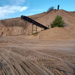 Kopalnia odkrywkowa z hałdą i taśmociągiem, ślady opon na piaszczystym podłożu.