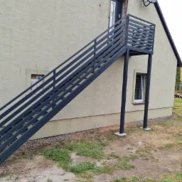 Zewnętrzne, metalowe schody z poziomymi szczeblami prowadzące do drzwi na piętrze budynku z beżową elewacją.