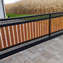 Nowoczesna brama przesuwna z czarną ramą ozdobioną motywem greckim i wypełnieniem z pionowych, drewnopodobnych paneli, na tle pola kukurydzy i kostki brukowej.