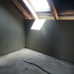 Wnętrze poddasza po nałożeniu gładzi cementowej na ściany i podłogę, widoczne okno dachowe i drewniane elementy konstrukcyjne, kabel elektryczny na podłodze.