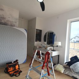 Dominik Marciniak - Czarny wentylator sufitowy z włączonym światłem, zamontowany w jasnym pokoju. Widoczna drabina, torba z narzędziami i narzędzia na podłodze. Ukończony montaż oświetlenia.