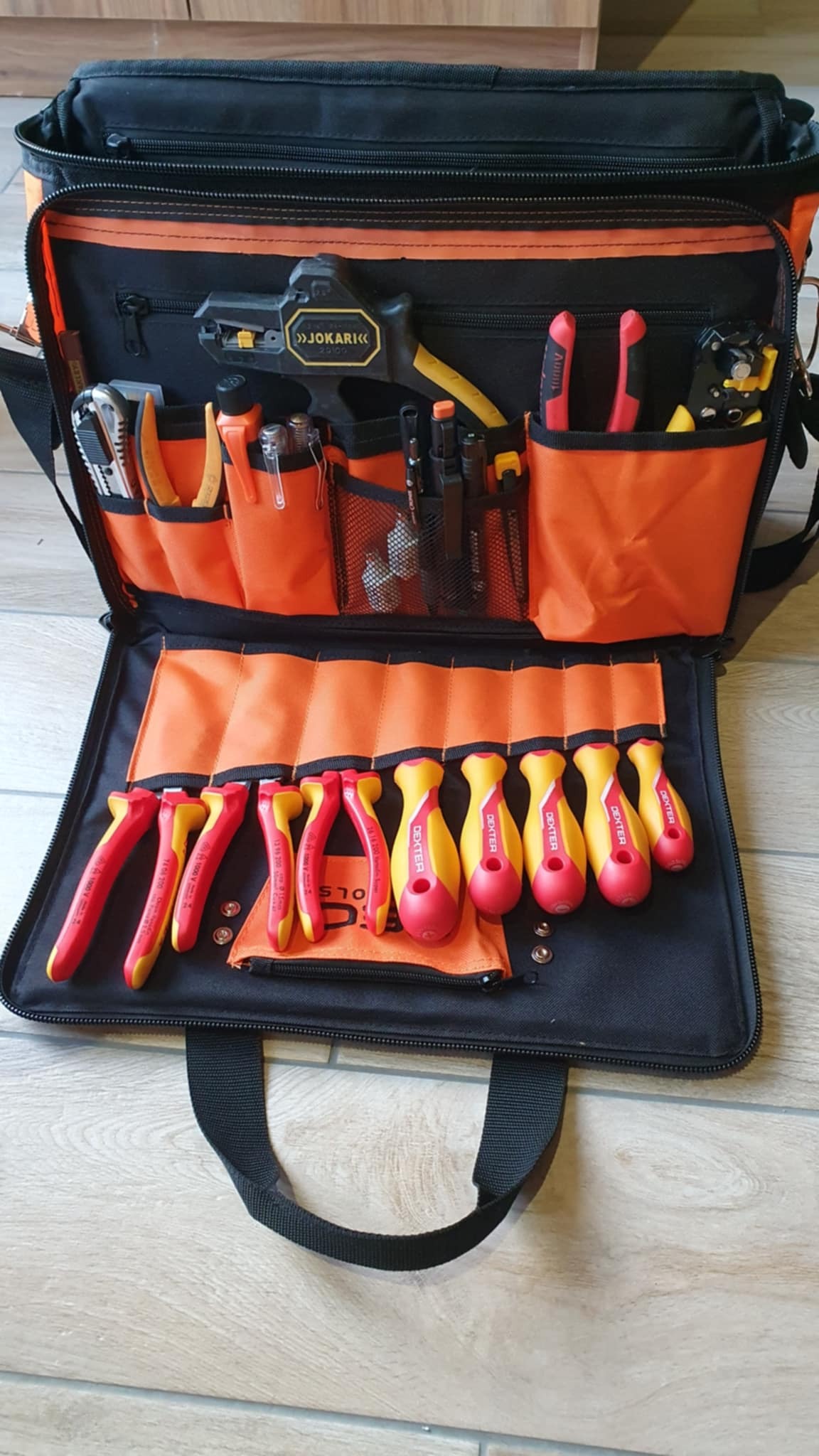 Otwarta torba narzędziowa z pomarańczowym wnętrzem, zawierająca zestaw izolowanych narzędzi: szczypce, wkrętaki. Widoczny ściągacz izolacji JOKARI.
