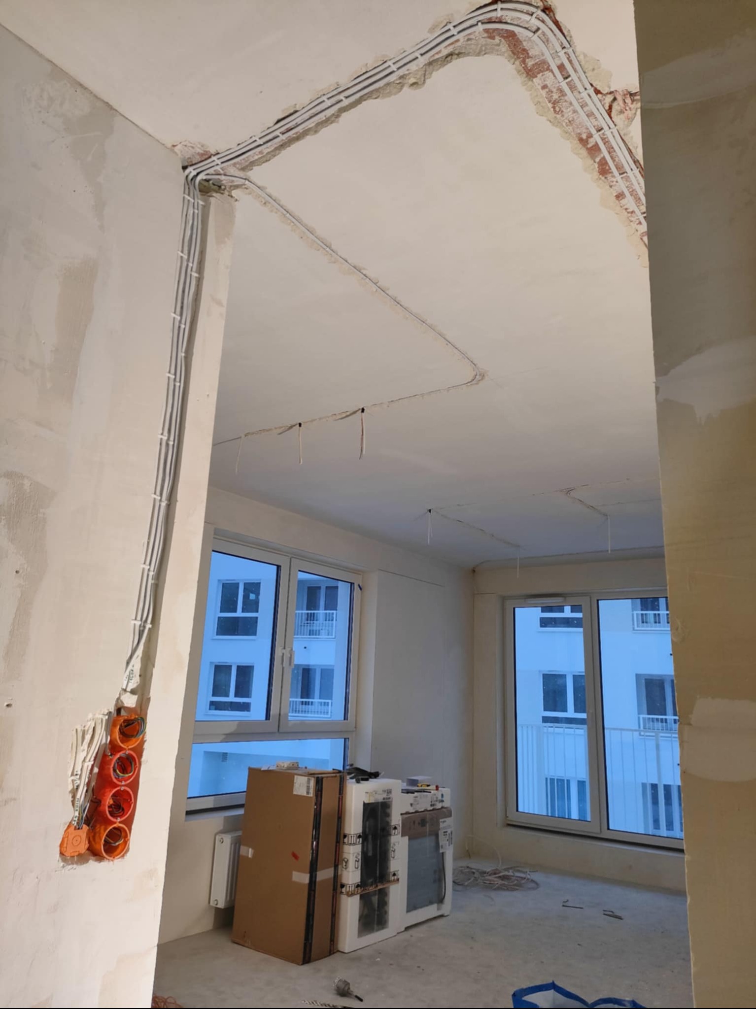 Wnętrze w trakcie instalacji elektrycznej: kable prowadzone po ścianach i suficie, puszki elektryczne, okna z widokiem na budynek.