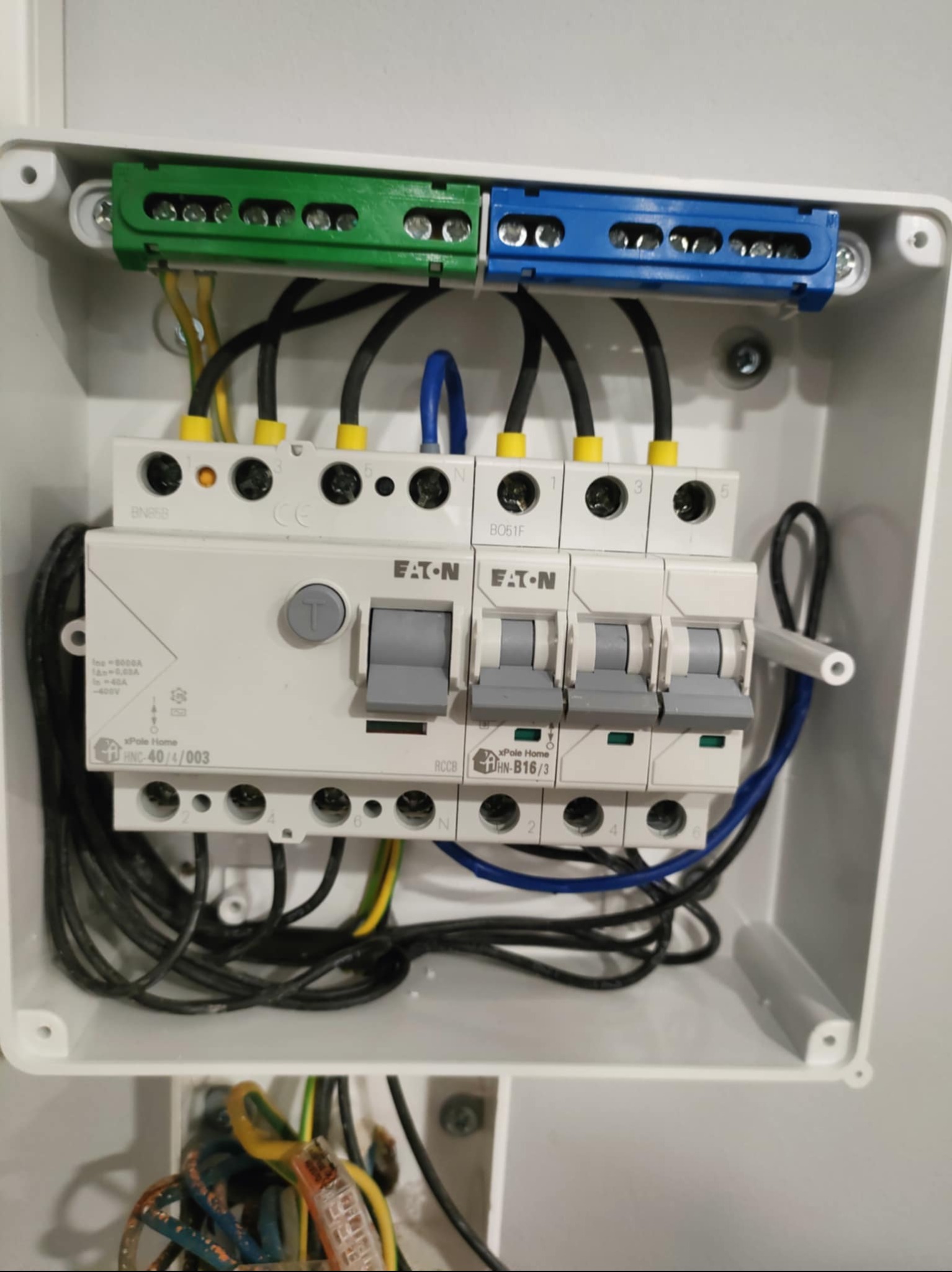 Rozdzielnica elektryczna Eaton w białej obudowie, widoczne zabezpieczenia, przewody w kolorach: czarnym, niebieskim, żółto-zielonym. Złącza zielone i niebieskie.
