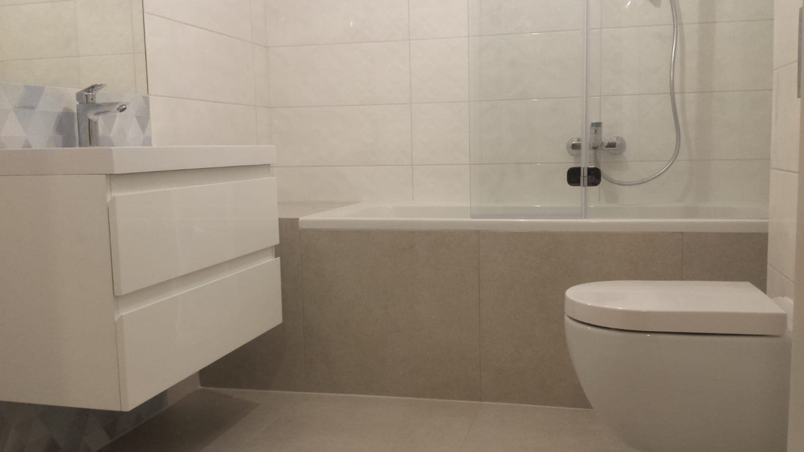 Nowoczesna łazienka z białymi płytkami, umywalką z szafką, wanną z parawanem i podwieszaną toaletą.