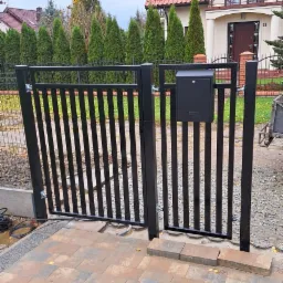 Czarna furtka z pionowymi szczebelkami i skrzynką na listy, prowadząca do posesji z kostki brukowej, w tle dom z czerwoną dachówką i rząd zielonych drzew.