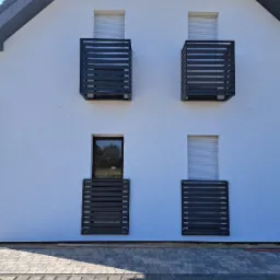 Elewacja domu z białego tynku z czterema nowoczesnymi, metalowymi balustradami okiennymi w kolorze antracytowym, widok z zewnątrz na kostkę brukową.