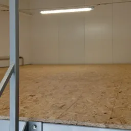 Wnętrze magazynu z podwyższoną podłogą z płyty OSB i metalową konstrukcją wsporczą, oświetlone lampą jarzeniową.