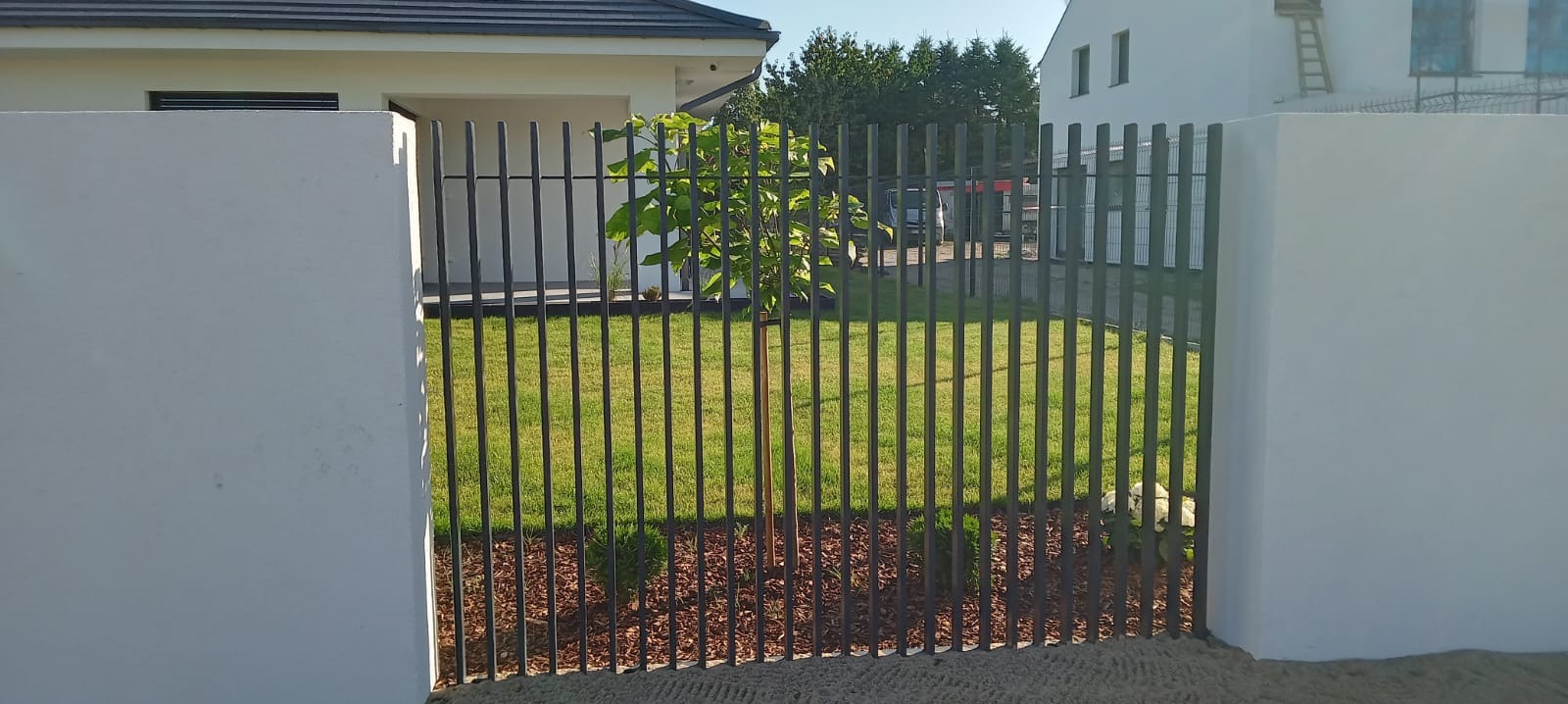 Nowoczesne ogrodzenie z pionowych, grafitowych paneli między białymi, murowanymi słupkami, z młodym drzewkiem zasadzonym wewnątrz posesji, widoczne budynki w tle.