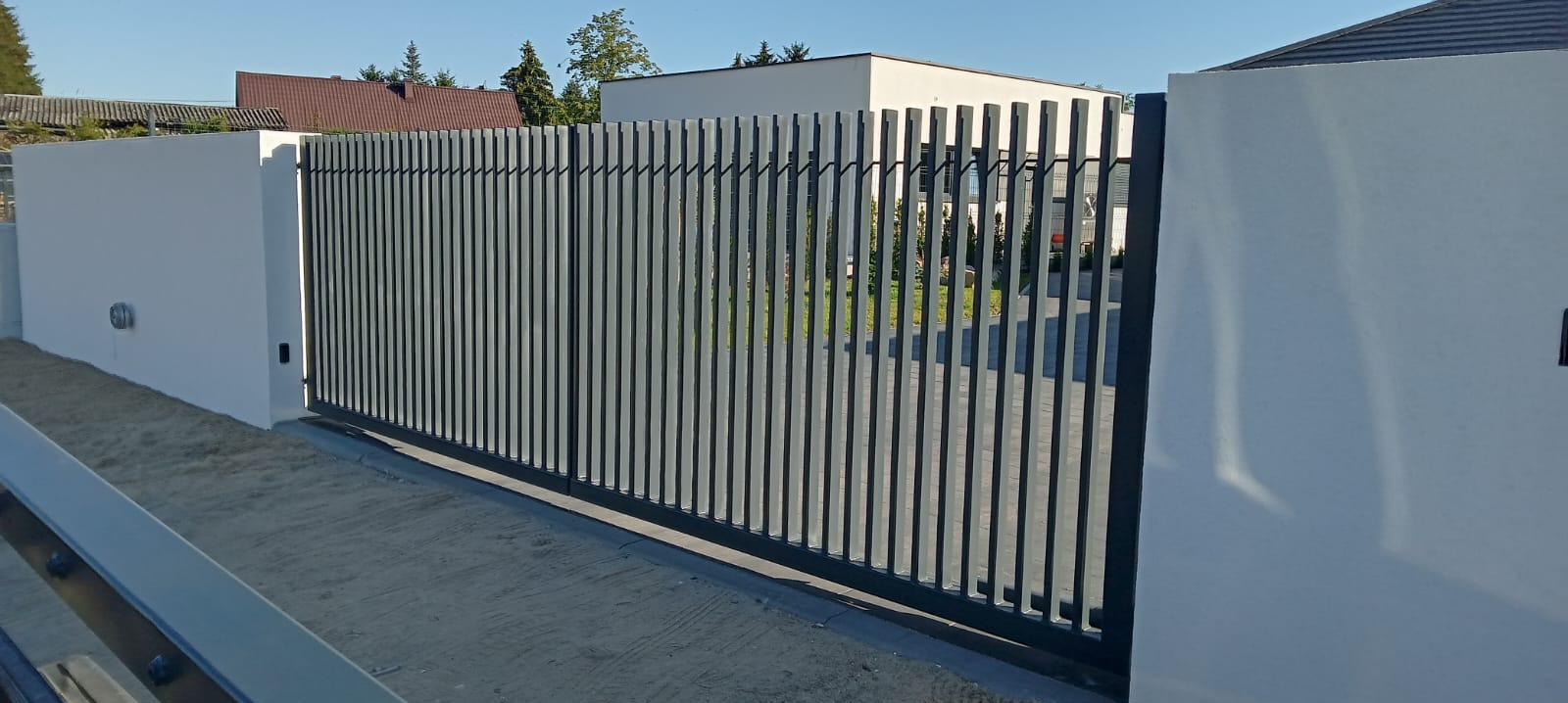 Nowoczesna brama przesuwna z pionowych profili, zamontowana między dwoma białymi słupkami, z widokiem na dom w tle.