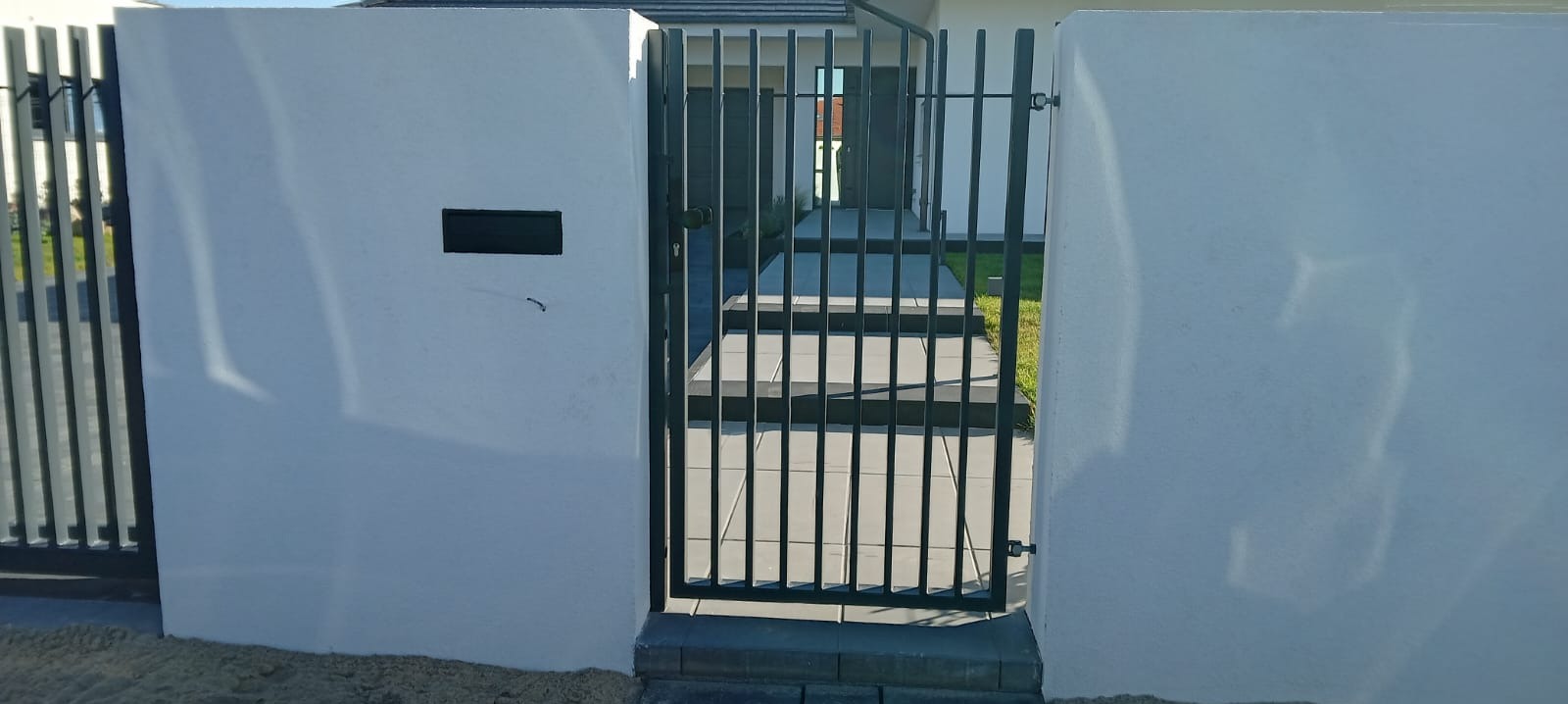 Nowoczesna furtka z metalowymi prętami, wkomponowana w białe, murowane słupki z zamontowaną skrzynką na listy, prowadząca do domu z szarymi schodami.