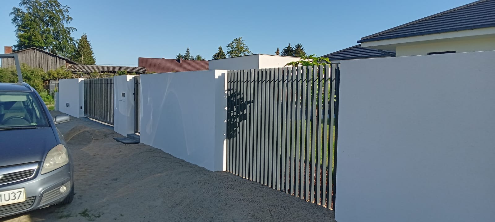 Nowoczesne ogrodzenie z białego muru i pionowych, metalowych paneli w kolorze grafitowym, z bramą wjazdową w tle, na piaszczystym podłożu, z samochodem osobowym zaparkowanym z boku.