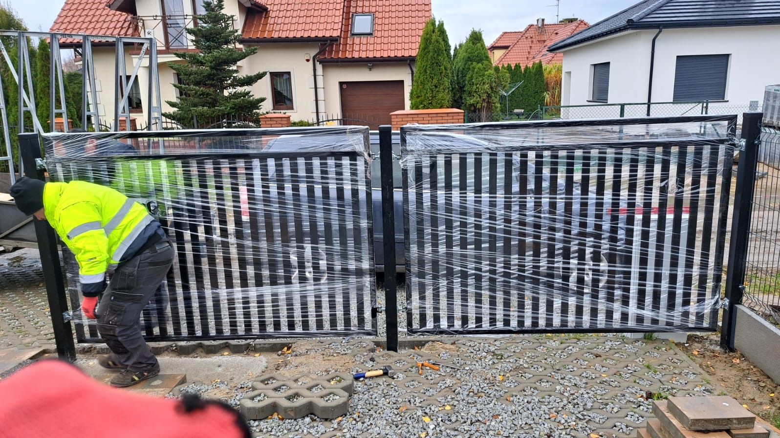 Montaż czarnej, pionowo żebrowanej bramy wjazdowej, zabezpieczonej folią, przez pracownika w odblaskowej kurtce na posesji z kostką ażurową i domami w tle.