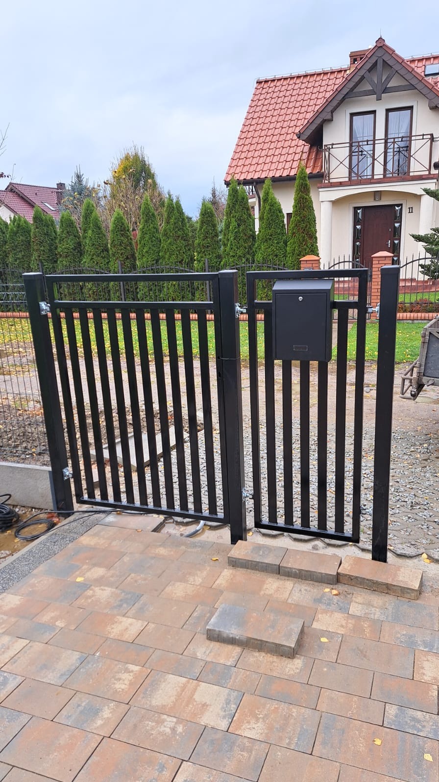 Czarna furtka z pionowymi szczebelkami i skrzynką na listy, prowadząca do posesji z kostki brukowej, w tle dom z czerwoną dachówką i rząd zielonych drzew.