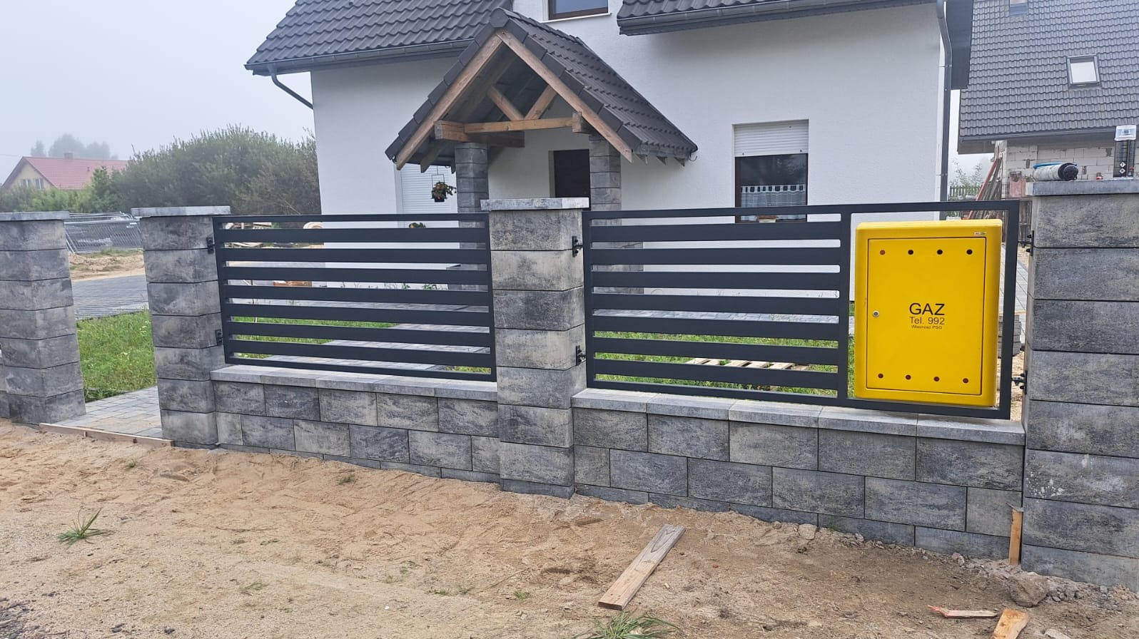 Nowoczesne ogrodzenie panelowe z szarej kostki i poziomych, ciemnych przęseł, z żółtą skrzynką gazową po prawej stronie, na tle domu w mglisty dzień.