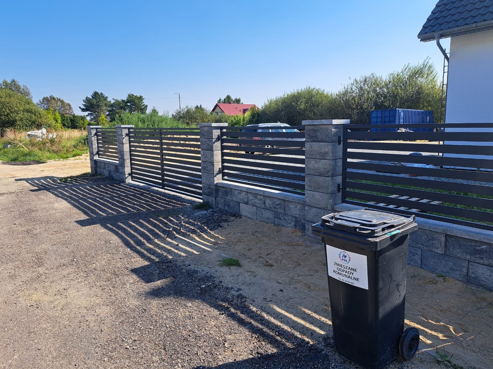 Nowoczesne ogrodzenie posesji z poziomych paneli stalowych i słupków z szarej kostki, widoczne z zewnątrz, z czarnym pojemnikiem na odpady komunalne w pierwszym planie.