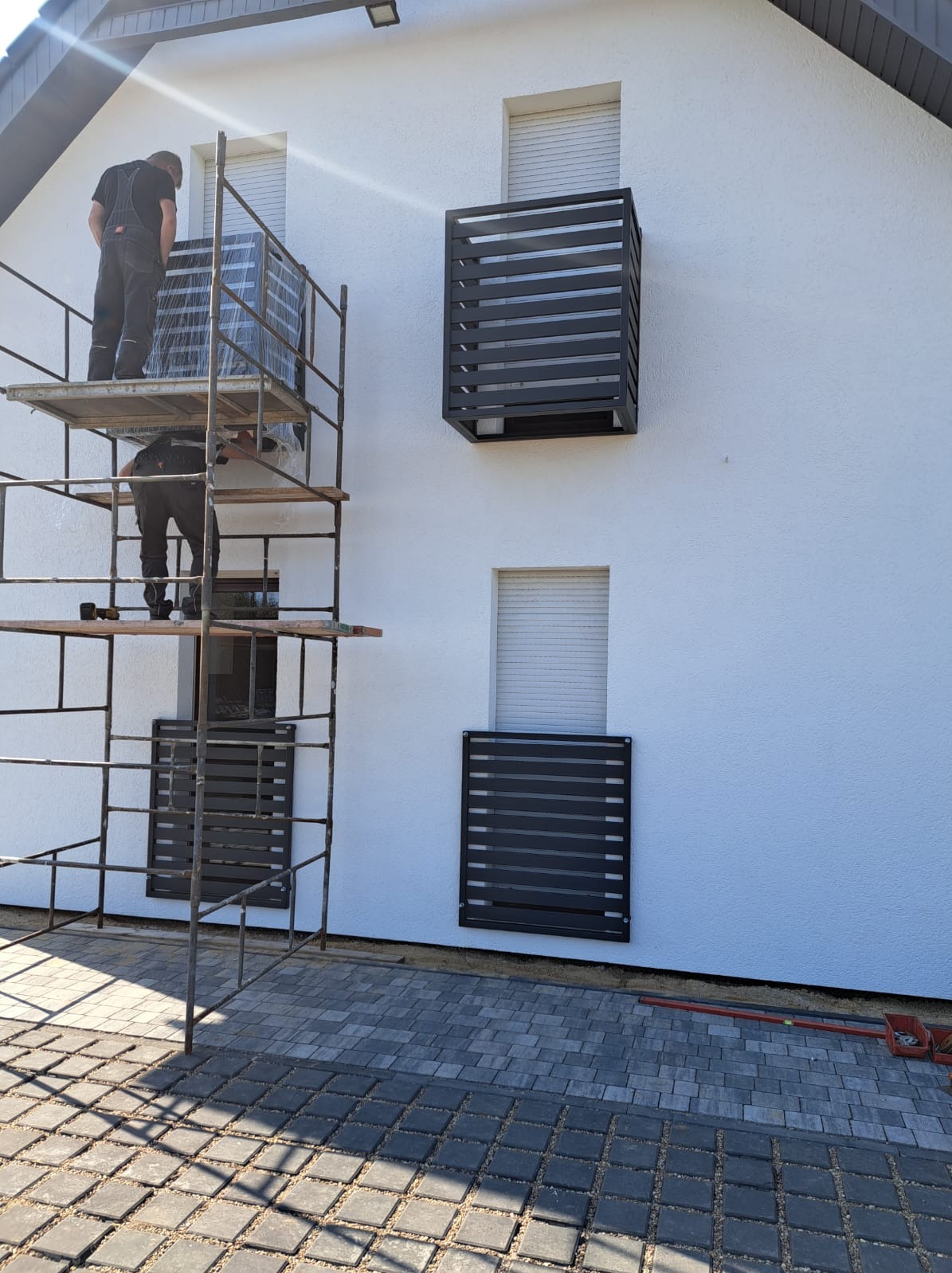 Montaż metalowych osłon okiennych na elewacji domu przez dwóch pracowników na rusztowaniu. Widoczne okna z roletami i kostka brukowa na podwórku.
