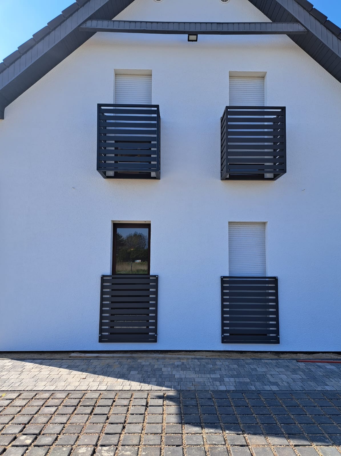 Elewacja domu z białego tynku z czterema nowoczesnymi, metalowymi balustradami okiennymi w kolorze antracytowym, widok z zewnątrz na kostkę brukową.