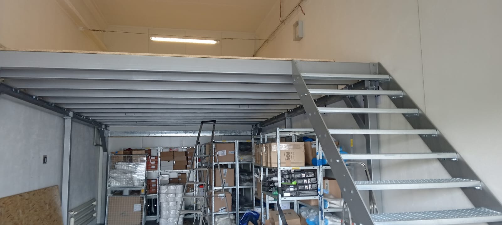 Metalowa konstrukcja antresoli z schodami w magazynie, widoczne półki z kartonami i drabina.