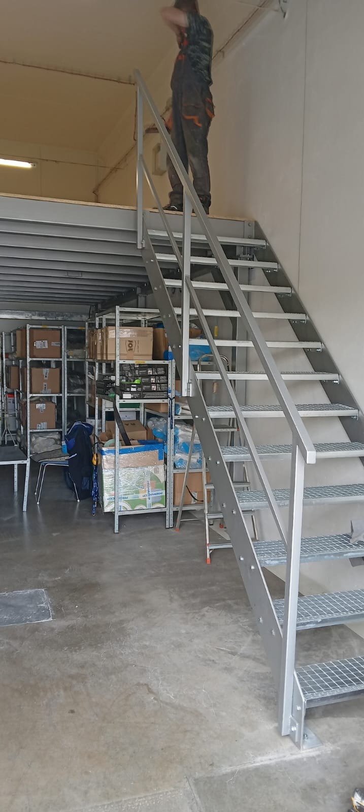 Metalowe schody z kratowymi stopniami i poręczą, prowadzące na antresolę magazynu, z pracownikiem w kombinezonie na górze.