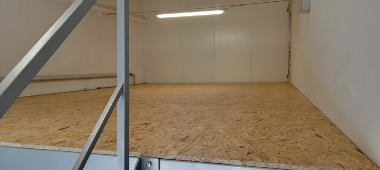 Wnętrze magazynu z podwyższoną podłogą z płyty OSB i metalową konstrukcją wsporczą, oświetlone lampą jarzeniową.