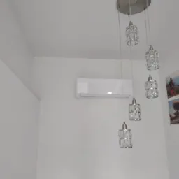 Biała jednostka klimatyzacji ściennej w pokoju z białymi ścianami, obok wiszącej lampy z kryształkami i obrazków.