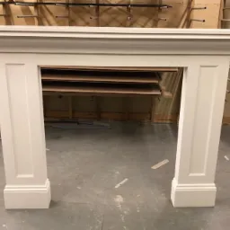 Biały, klasyczny portal kominkowy w trakcie produkcji, widoczne niedoskonałości wykończenia i elementy warsztatu w tle.