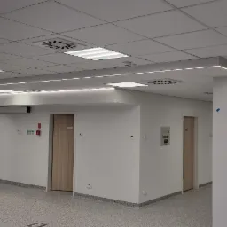 Nowoczesne wnętrze biurowe z podwieszanym sufitem, oświetleniem LED wzdłuż filarów i jasnymi drzwiami.