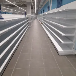 Wnętrze pustego sklepu z szarymi płytkami podłogowymi i rzędami białych regałów sklepowych pod niebieską ścianą z metalowymi kratkami, oświetlone długimi lampami sufitowymi.