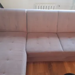 Jasnoróżowa, welurowa kanapa narożna z widocznymi guzikami na siedzisku i oparciu, stojąca na parkiecie, z fragmentem drabiny i grzejnika w tle.