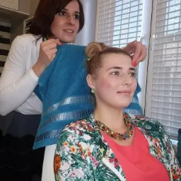 Klientka w kwiecistej kurtce podczas przygotowań do zabiegu kosmetycznego, z upiętymi włosami i kolorowym naszyjnikiem, trzymana przez kosmetyczkę za pomocą niebieskiego ręcznika.