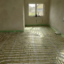 Wnętrze w trakcie instalacji ogrzewania podłogowego: rury grzewcze ułożone na folii z siatką, widoczne okna i surowe ściany.