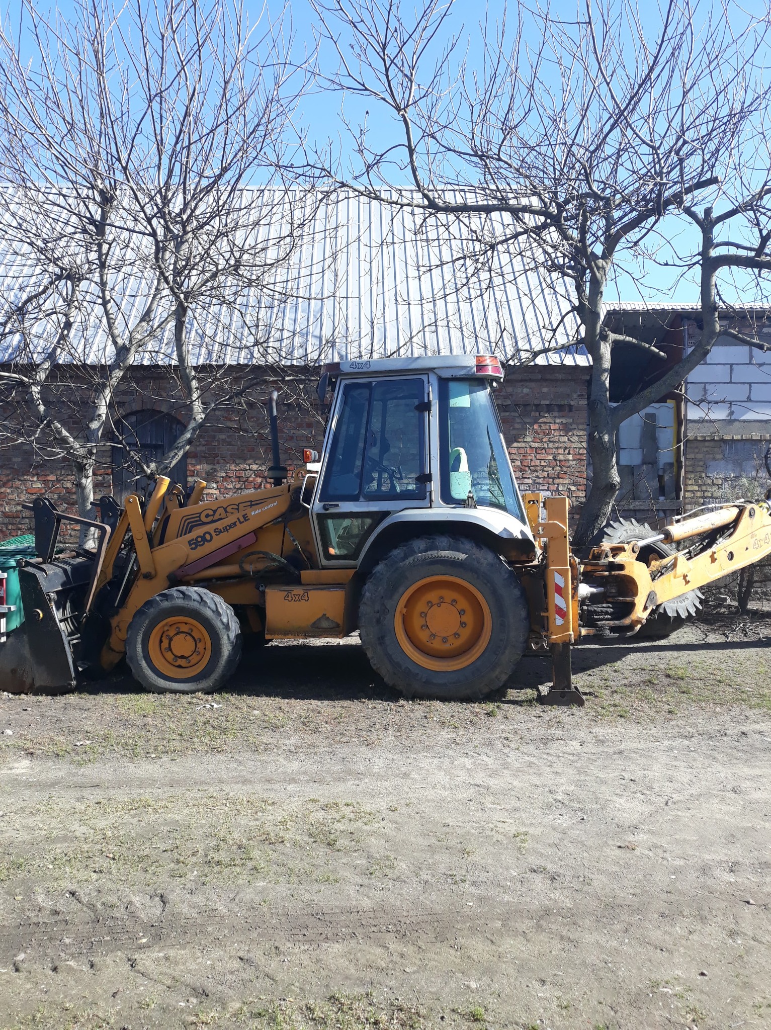Żółto-szara koparko-ładowarka Case 590 Super LE zaparkowana na gruntowej powierzchni, widoczne drzewa bez liści i budynek z cegły w tle.