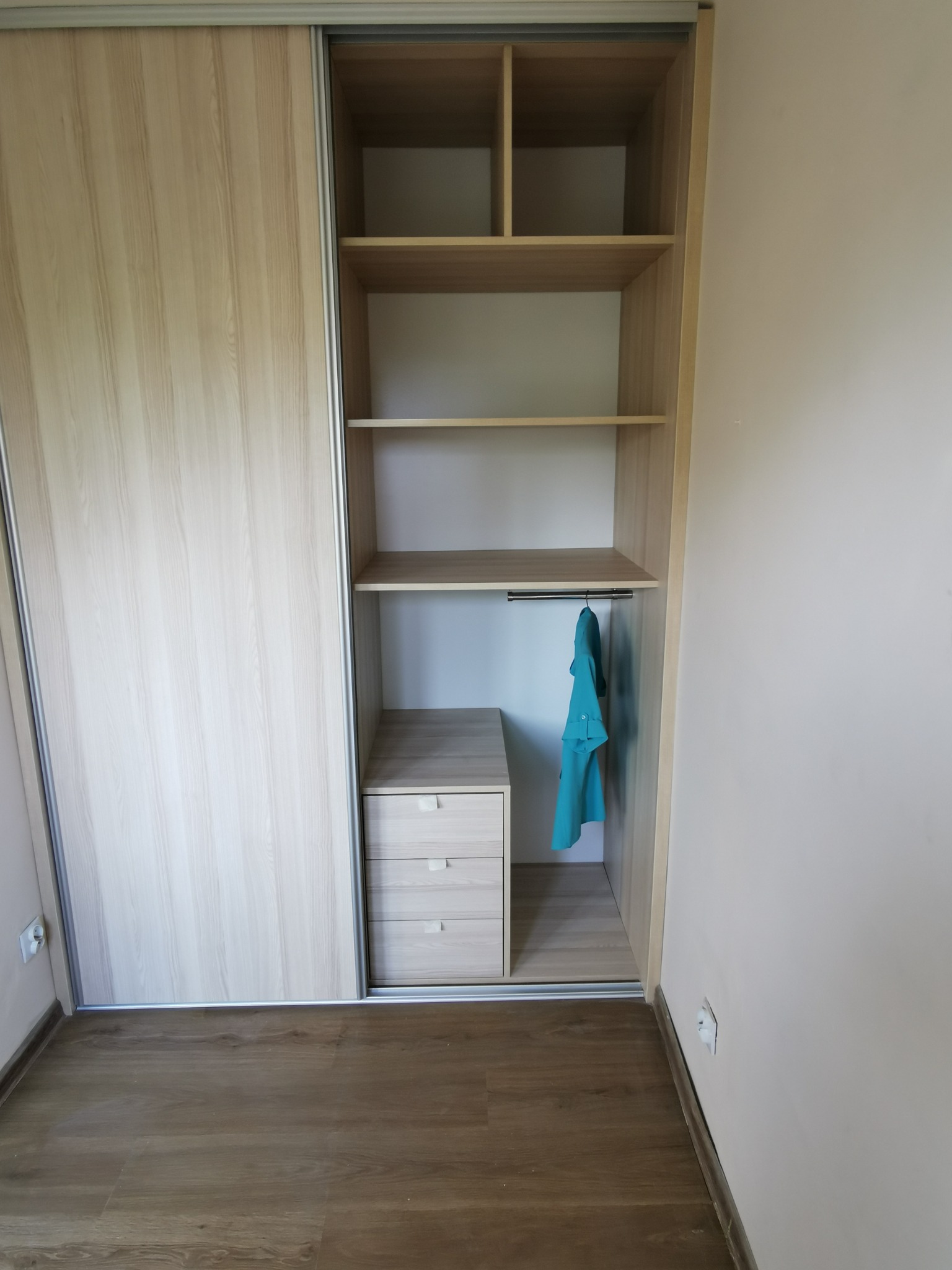 Jasna szafa wnękowa z przesuwnymi drzwiami, otwartymi półkami i trzema szufladami, z błękitną koszulą wiszącą na wieszaku.