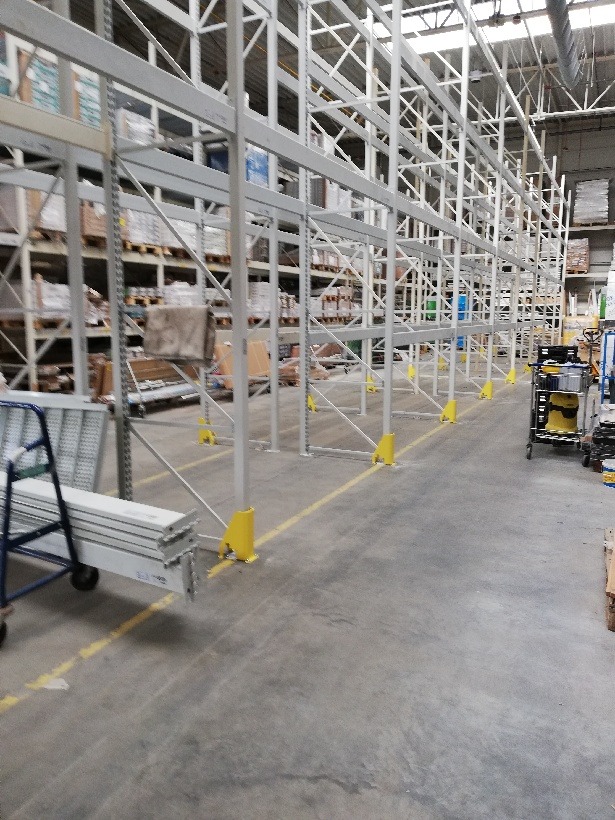 Wysokie, białe regały magazynowe z żółtymi zabezpieczeniami w magazynie firmy z Warszawy, wypełnione różnorodnymi towarami.