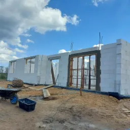 Budowa domu murowanego z betonu komórkowego w trakcie wznoszenia ścian, widoczne zbrojenia, drewniane stemple podtrzymujące nadproża oraz taczka i pojemnik budowlany na placu budowy.